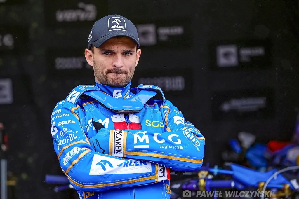 Zawodnik żużlowy w niebieskim kombinezonie sportowym z założonymi rękami, na tle warsztatu i sprzętu sportowego.