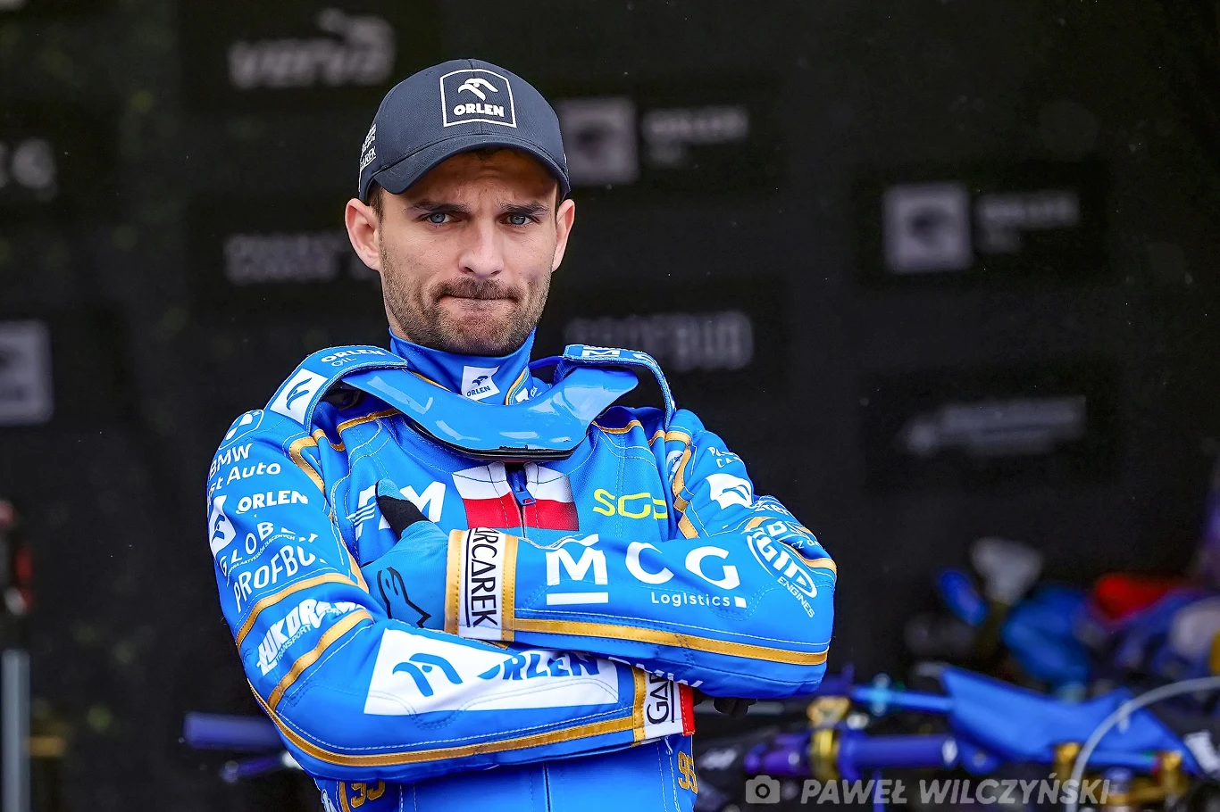 Bartosz Zmarzlik Zawodnik żużlowy w niebieskim kombinezonie sportowym z założonymi rękami, na tle warsztatu i sprzętu sportowego.