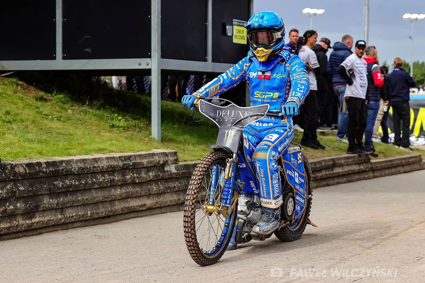 Zawodnik w niebieskim kombinezonie i kasku jedzie motocyklem żużlowym po torze, w tle stoi grupa osób obserwujących wydarzenie.