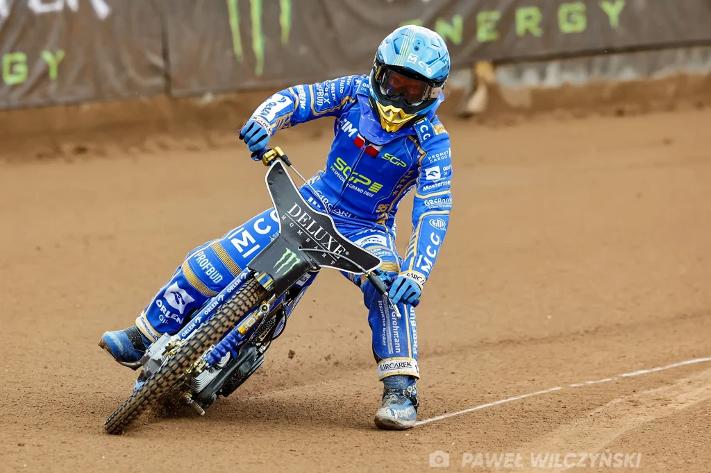 Bartosz Zmarzlik w Vojens Zawodnik żużlowy w niebieskim kombinezonie na motocyklu podczas wyścigu na torze żużlowym, intensywna rywalizacja, dynamiczna pozycja sportowca, widoczne detale stroju i motocykla wraz z licznymi sponsorami.