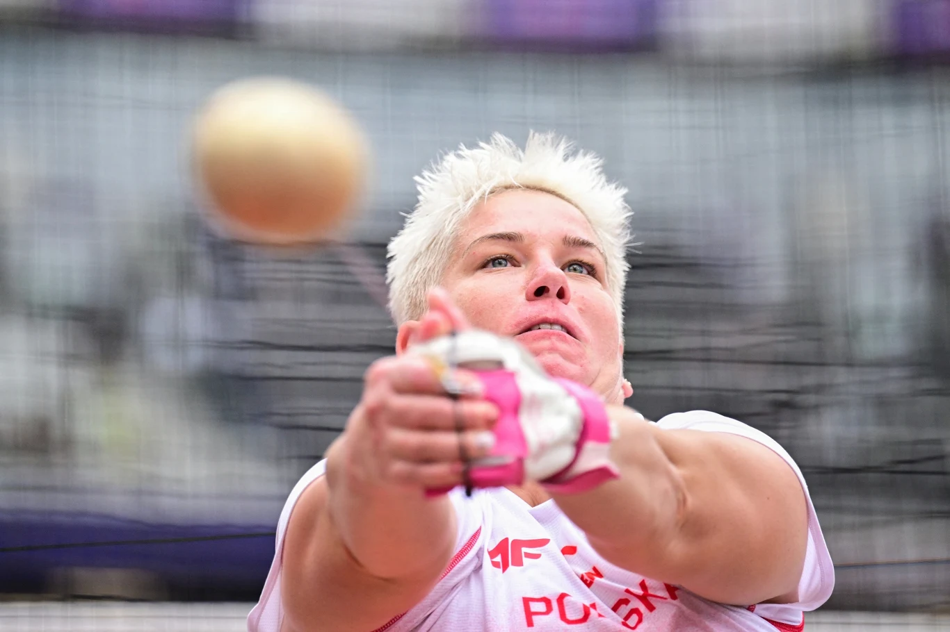 Zbliżenie na skupioną zawodniczkę podczas rzutu młotem na stadionie, wyraźnie widoczny młot w ruchu oraz determinacja na twarzy sportowczyni.