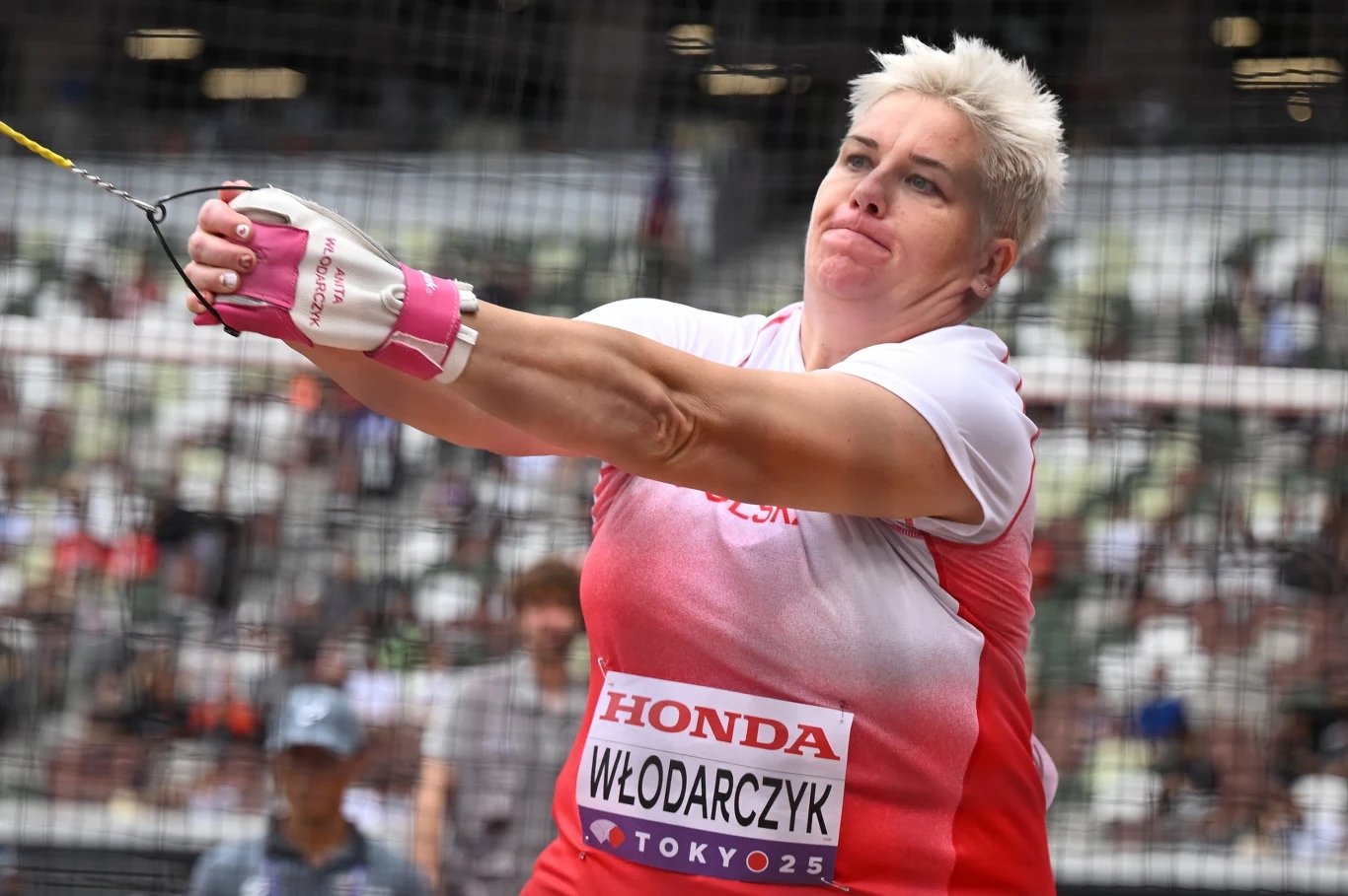 Zawodniczka podczas rzutu młotem na stadionie lekkoatletycznym, ujęcie skupione na dynamicznej postawie oraz wyraźnie widocznych emocjach sportowca, w tle zamazana publiczność.