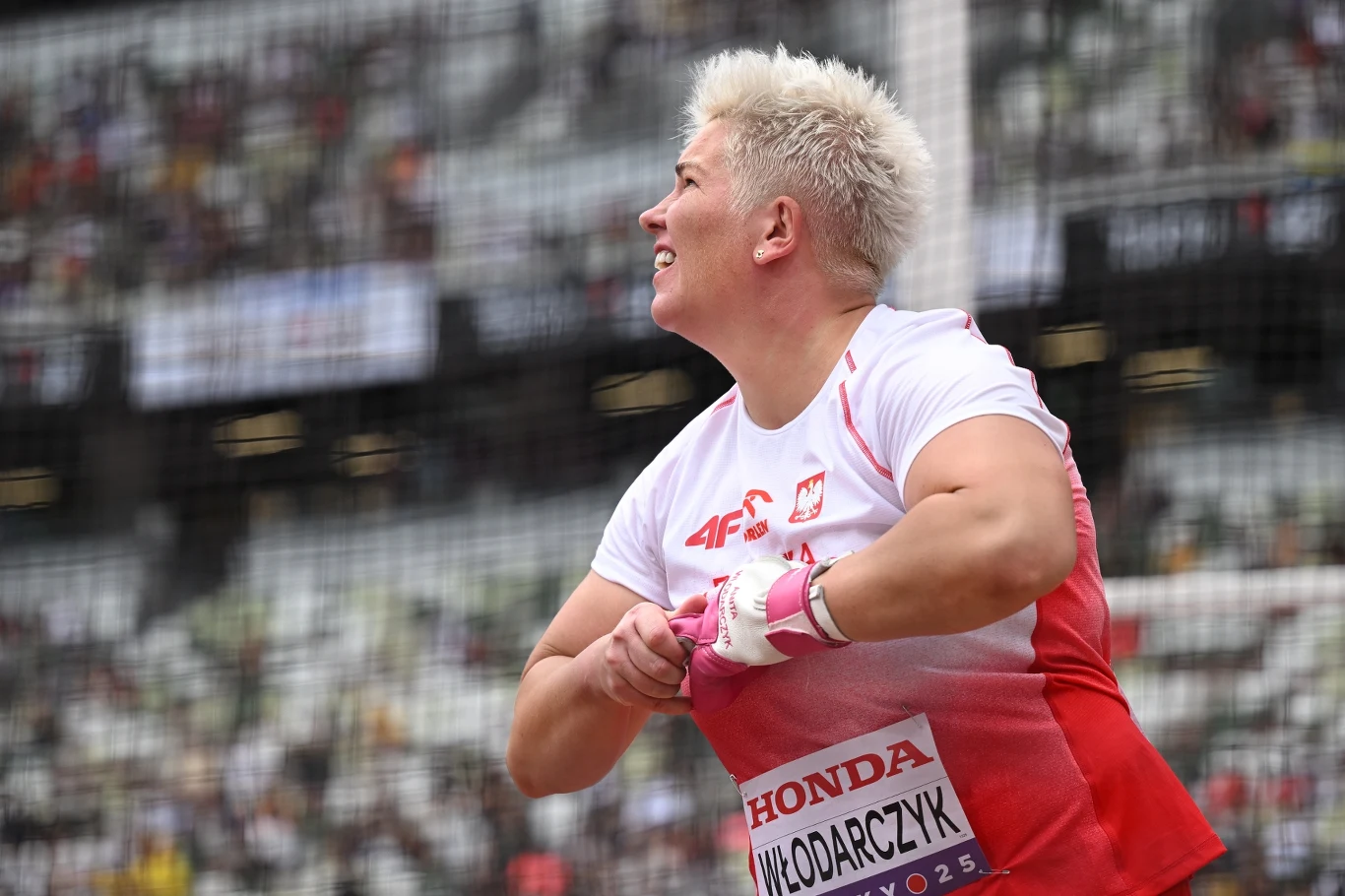 Zawodniczka w stroju sportowym z polską flagą i nazwiskiem Włodarczyk na piersi, przygotowująca się do rzutu młotem na stadionie lekkoatletycznym, w tle widoczna rozmyta publiczność i siatka ochronna.