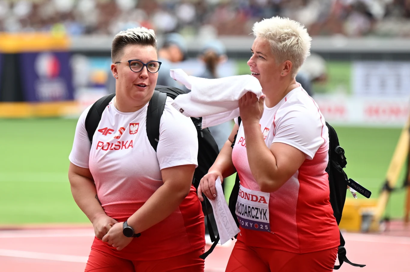 Dwie polskie lekkoatletki w strojach reprezentacji Polski, uśmiechnięte, spacerujące po stadionie lekkoatletycznym, z torbami sportowymi na ramionach.
