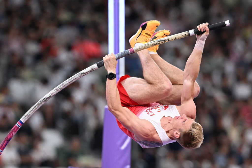 Lekkoatleta w biało-czerwonym stroju podczas skoku o tyczce, dynamiczny moment w locie z napiętymi mięśniami, na tle zamazanej publiczności stadionowej.