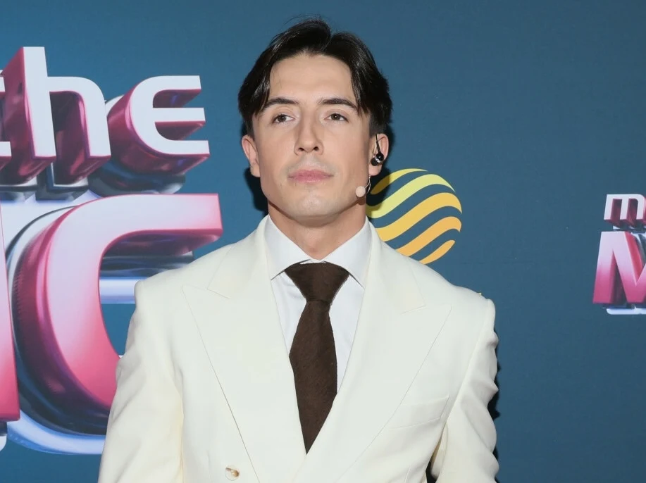 Dawid Kwiatkowski otworzył się przed fanami Elegancko ubrany mężczyzna w białym garniturze i brązowym krawacie stoi na tle ścianki z napisem, nosi zestaw słuchawkowy.