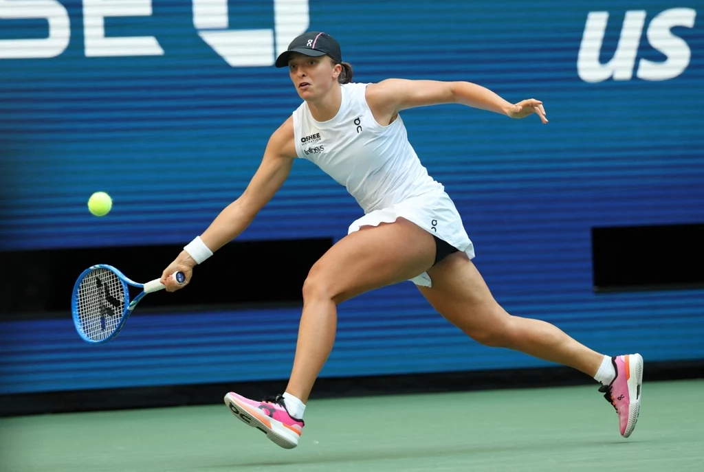 Tenisistka w białym stroju sportowym dynamicznie odbija piłkę na korcie podczas meczu, tło stanowi niebieska tablica z napisem US Open.