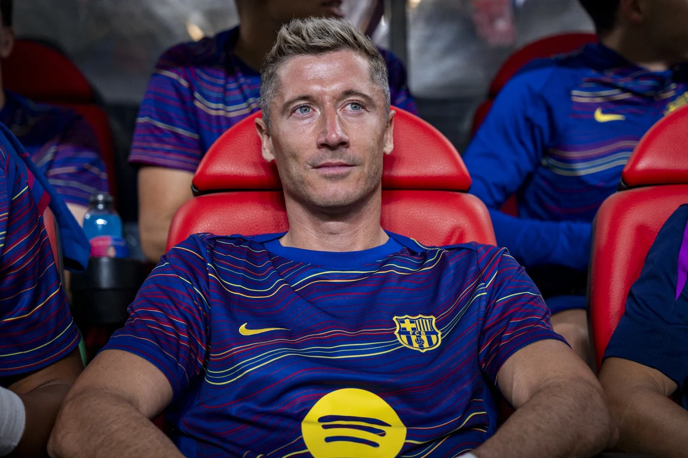 Piłkarz w koszulce FC Barcelony siedzi na ławce rezerwowych, skupiony, w tle widoczni inni zawodnicy zespołu w podobnych strojach sportowych.