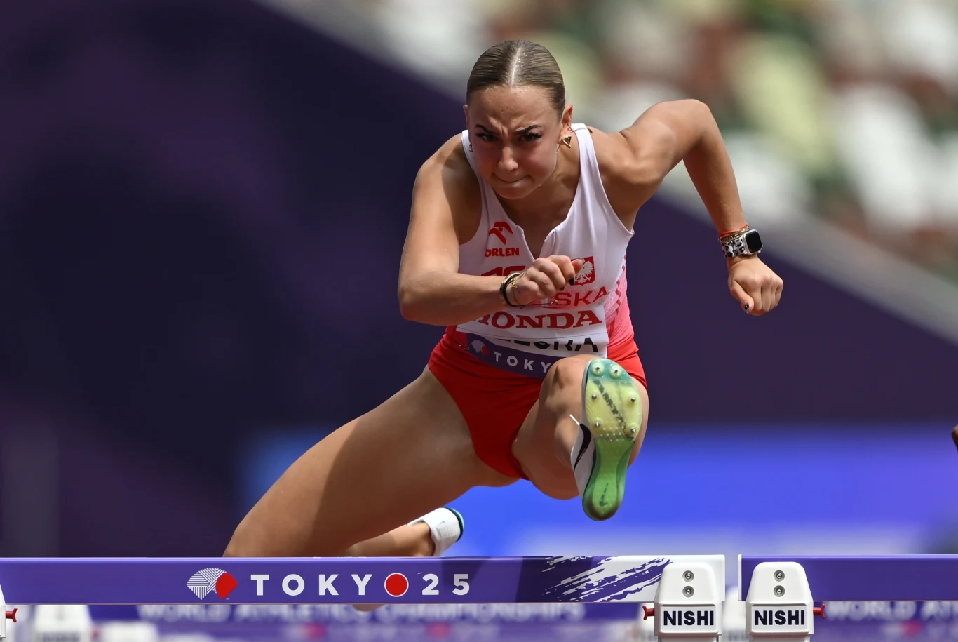 Lekkoatletka w stroju reprezentacji Polski wykonuje dynamiczny skok przez płotek podczas zawodów lekkoatletycznych na igrzyskach olimpijskich w Tokio 2025, widoczna determinacja, profesjonalny strój sportowy i napięcie mięśni.