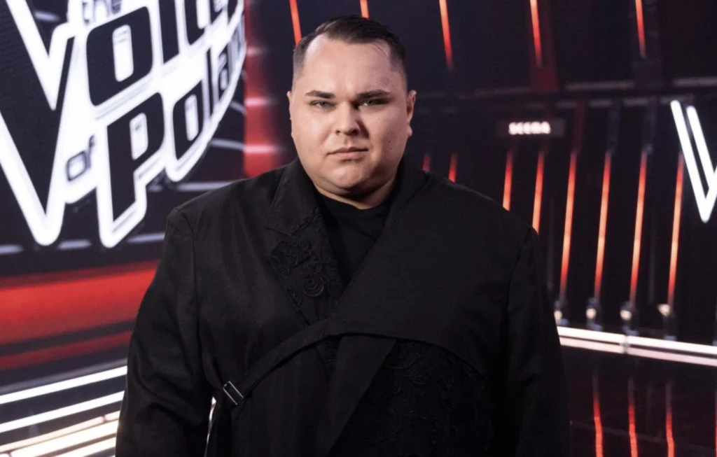 Mateusz Włodarczyk wrócił do programu po 11 latach Mężczyzna w czarnym stroju stoi na tle podświetlonego napisu The Voice of Poland, scena z elementami czerwieni i światłami LED.