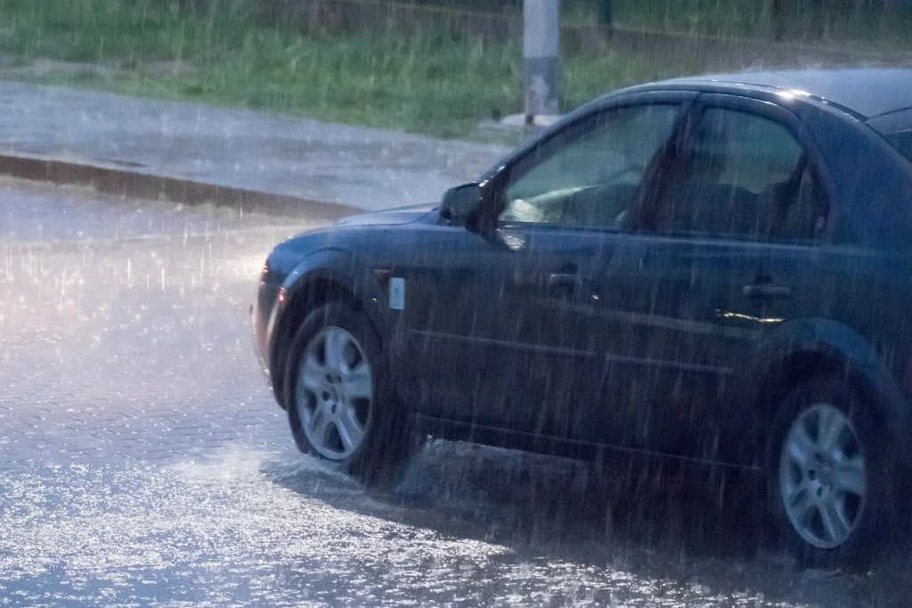 Samochód osobowy jadący ulicą podczas intensywnego deszczu, woda rozpryskuje się spod kół na mokrej nawierzchni.