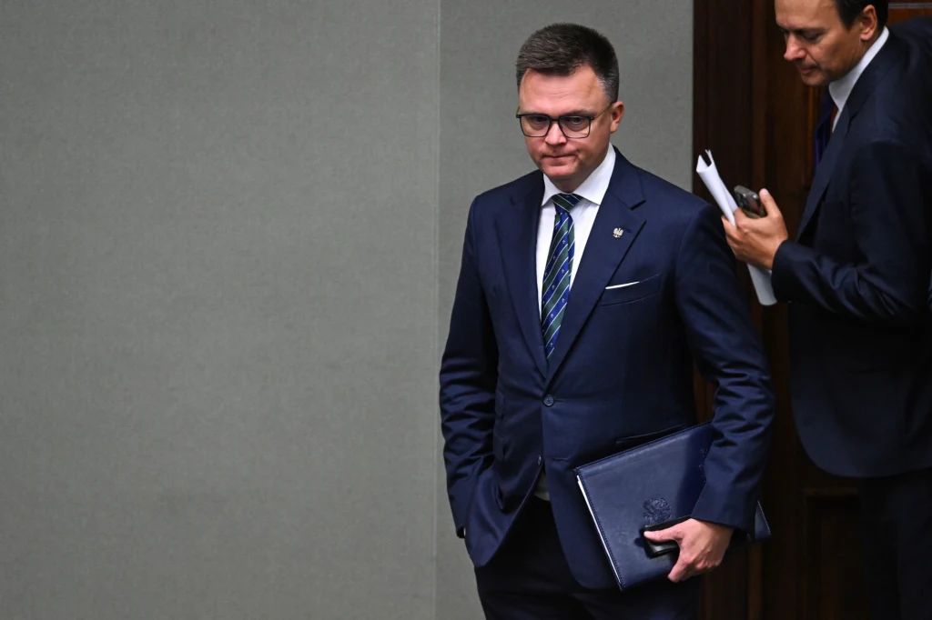 Mężczyzna w garniturze z aktówką w ręku stoi przy ścianie, w tle widoczna jest druga osoba z dokumentami.