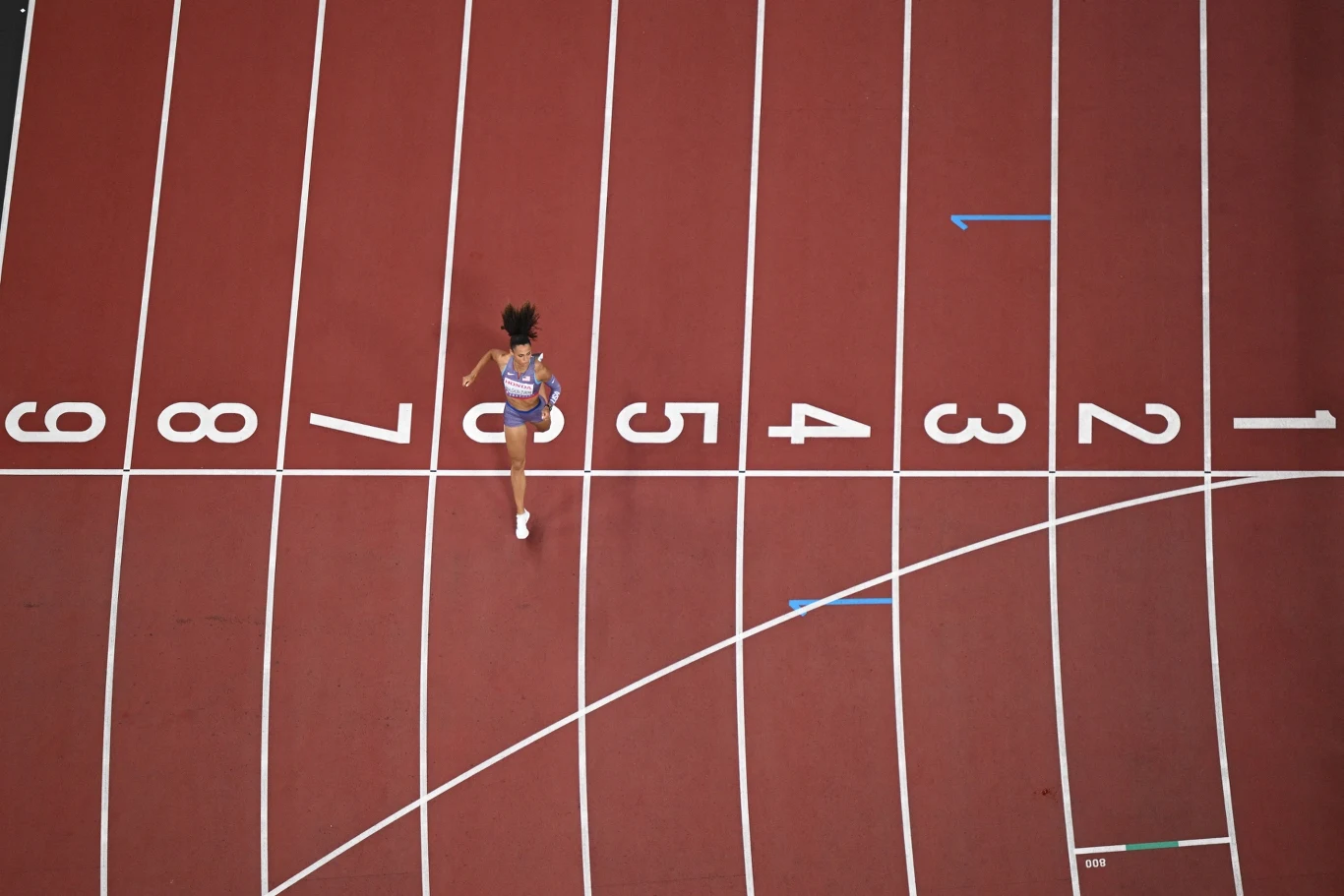 Zawodniczka biegnąca pojedynczo po pustym torze lekkoatletycznym, centralnie na torze oznaczonym numerem 5, otoczona liniami numeracji torów, widok z góry.