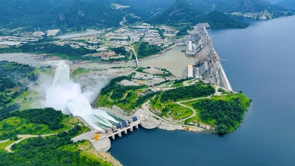 Zapora GERD / Wikimedia Commons Wielka zapora wodna na dużej rzece otoczona zielonymi wzgórzami, widoczne są spływające masy wody z tamy oraz infrastruktura hydrotechniczna i drogi wokół obiektu.