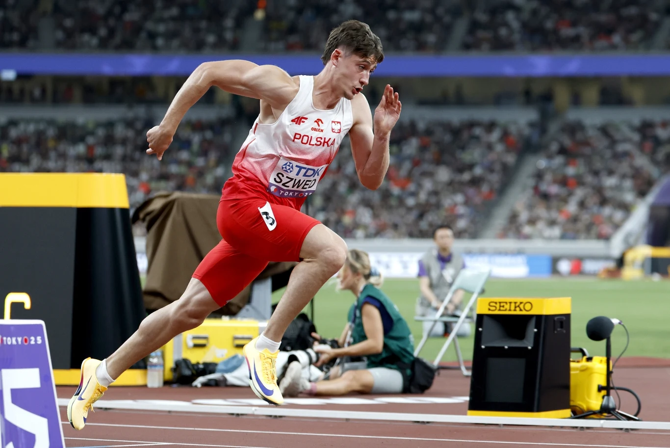 Lekkoatleta w stroju reprezentacji Polski rozpoczyna bieg na bieżni stadionu, w tle trybuny pełne kibiców oraz sprzęt sportowy.