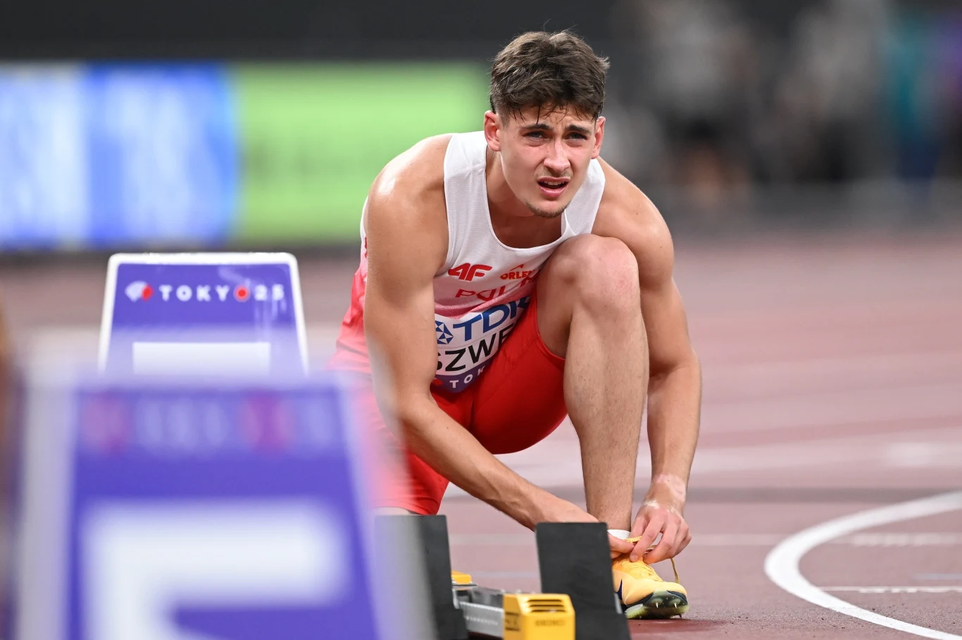 Lekkoatleta w białej i czerwonej koszulce przygotowuje się do biegu na stadionie lekkoatletycznym, klęcząc przy blokach startowych i wiążąc but, w tle tor bieżni i rozmyte elementy stadionu.