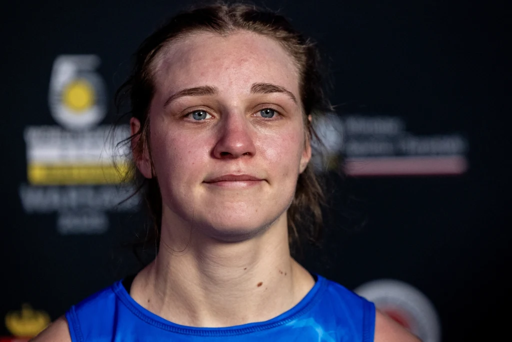 Julia Szeremeta Młoda kobieta w sportowym niebieskim stroju z lekkimi oznakami zmęczenia na twarzy stoi na tle ciemnej planszy z logo i napisami.