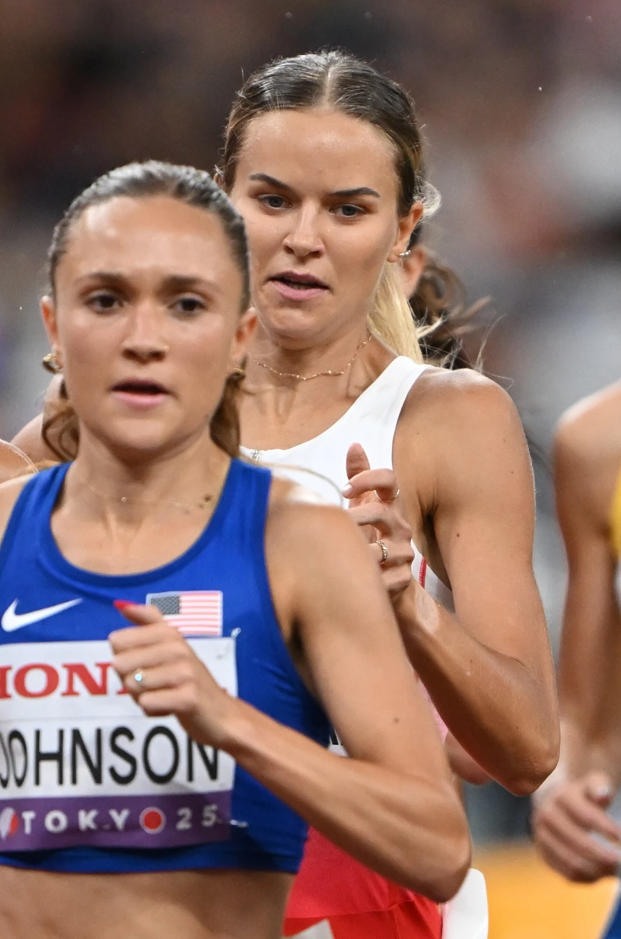 Dwie kobiety biegnące w wyścigu lekkoatletycznym na stadionie, widoczne z bliska; jedna z nich w stroju sportowym z napisem Johnson i emblematem USA, skupienie na twarzach i dynamika rywalizacji.
