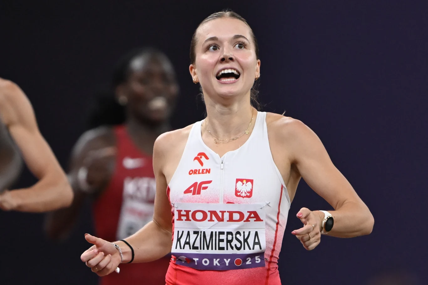 Polska lekkoatletka w stroju sportowym finiszuje podczas wyścigu na stadionie, z widocznym uśmiechem na twarzy, za nią zamazana sylwetka innej zawodniczki.