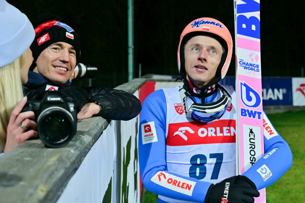 Dwóch mężczyzn przy barierce, jeden w stroju skoczka narciarskiego z nartą w ręku i numerem 87, drugi z aparatem fotograficznym, obaj uśmiechnięci, tło to bandy reklamowe i trawiaste podłoże.