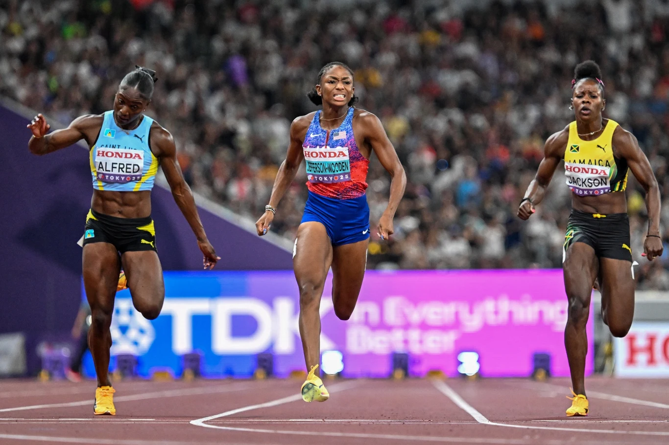 Trzy zawodniczki biegnące na ostatnich metrach wyścigu lekkoatletycznego na stadionie, każda skupiona na finiszu, w tle widownia oraz banery reklamowe.