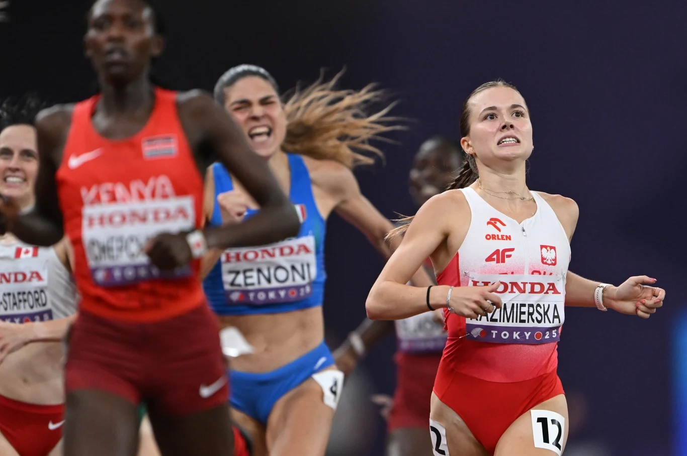 Kilka zawodniczek biegnących na stadionie podczas zawodów lekkoatletycznych, jedna z nich w biało-czerwonym stroju z napisem Polska wyraźnie wyprzedza pozostałe, twarze wyrażają determinację i emocje sportowej rywalizacji.