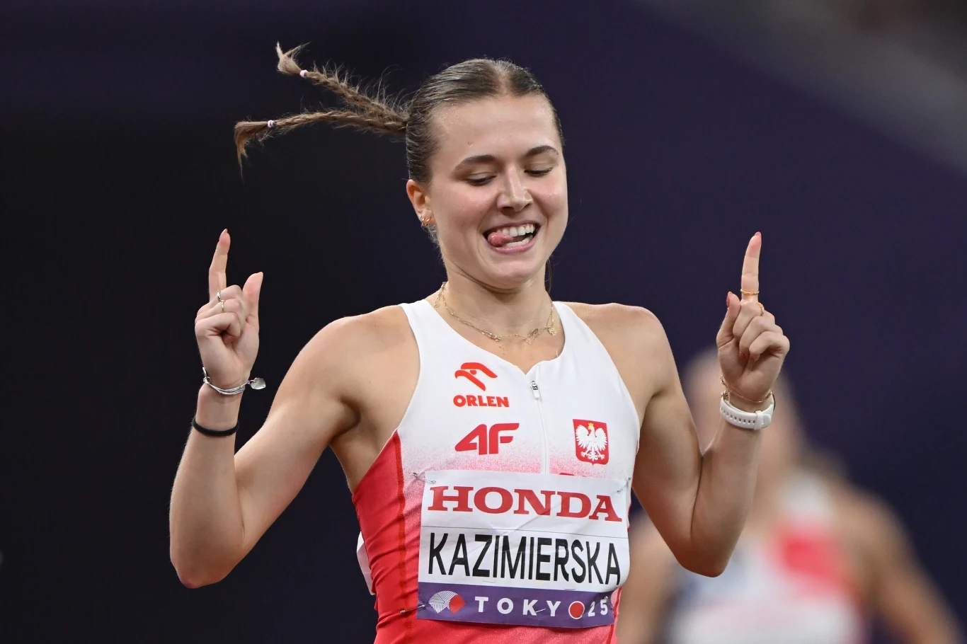 Zawodniczka lekkoatletyczna w stroju reprezentacji Polski z widocznym numerem startowym oraz nazwą sponsora, uśmiechnięta, z uniesionymi obiema dłońmi do góry na stadionie.