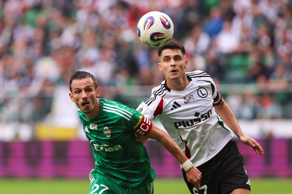 Legia Warszawa - Radomiak Radom Dwóch piłkarzy rywalizujących o piłkę w powietrzu podczas meczu, jeden w zielonym stroju z opaską kapitana, drugi w białym stroju z czarnymi elementami, w tle rozmyta publiczność na trybunach.