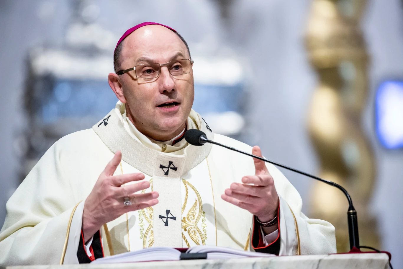 Prymas Polski abp Wojciech Polak Duchowny ubrany w białe szaty liturgiczne przemawia przy ambonie, mając przed sobą mikrofon i otwartą księgę, w tle rozmyte elementy wnętrza kościoła.