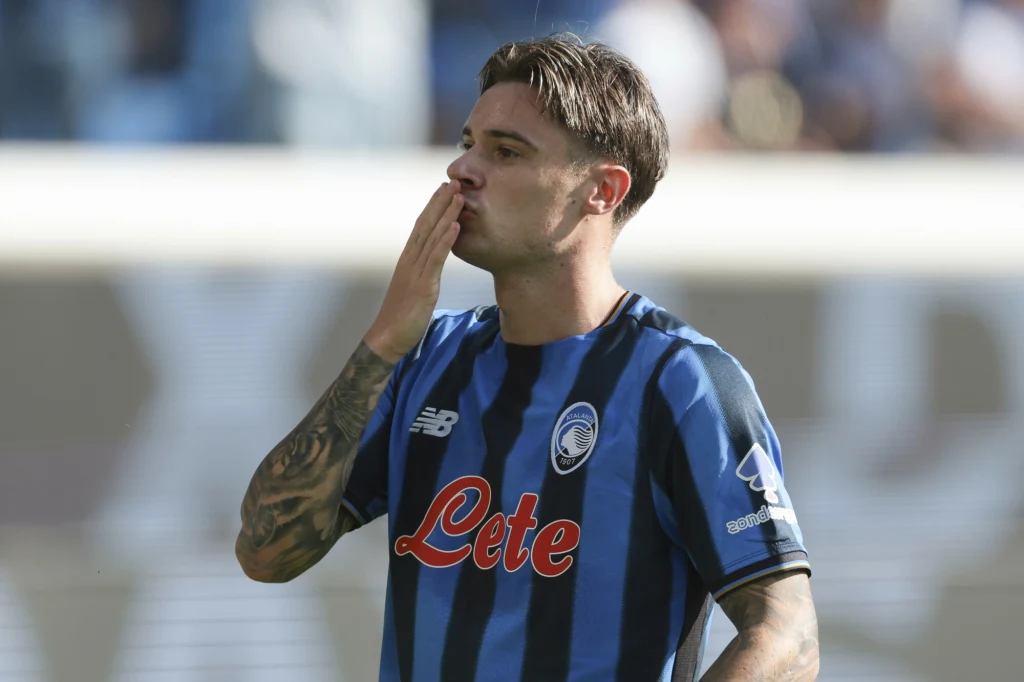 Nicola Zalewski Piłkarz w niebiesko-czarnej koszulce z logo klubu i charakterystycznym wzorem, wykonujący gest pocałunku skierowany w stronę trybun, na tle rozmytego stadionu.