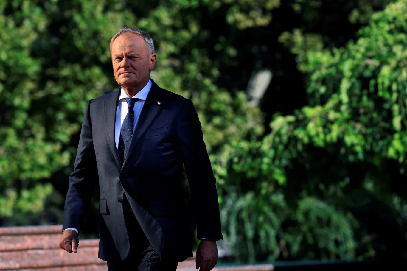 Premier Donald Tusk Mężczyzna w eleganckim garniturze idący na tle zielonych drzew, promienie słońca delikatnie oświetlają jego twarz, panuje poważny i dostojny nastrój.