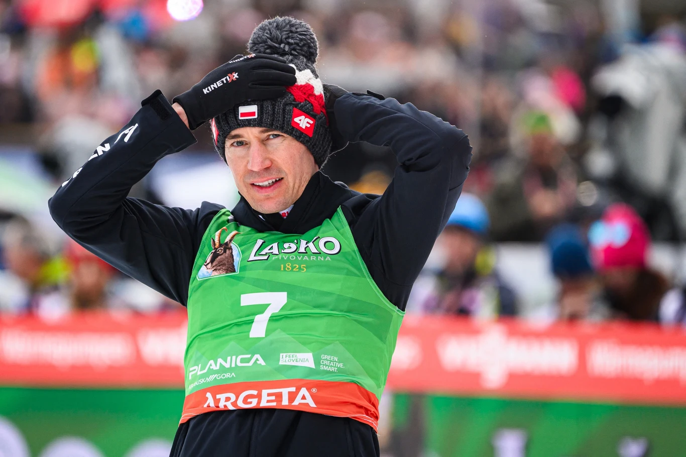 Sportowiec w zielonym numerze startowym oraz czapce na głowie, stojący na tle tłumu kibiców podczas zawodów narciarskich.