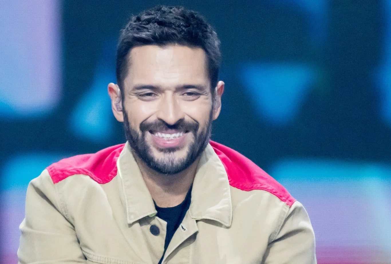 Mesajah wziął ślub Uśmiechnięty mężczyzna z krótkimi ciemnymi włosami i brodą ubrany w beżową kurtkę z czerwonymi wstawkami na ramionach na tle rozmytych, kolorowych świateł.