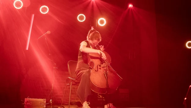 Muzyk grający na wiolonczeli siedzi na scenie oświetlonej ciepłym czerwonym światłem, wokół widoczne reflektory i elementy nagłośnienia tworzące koncertową atmosferę.