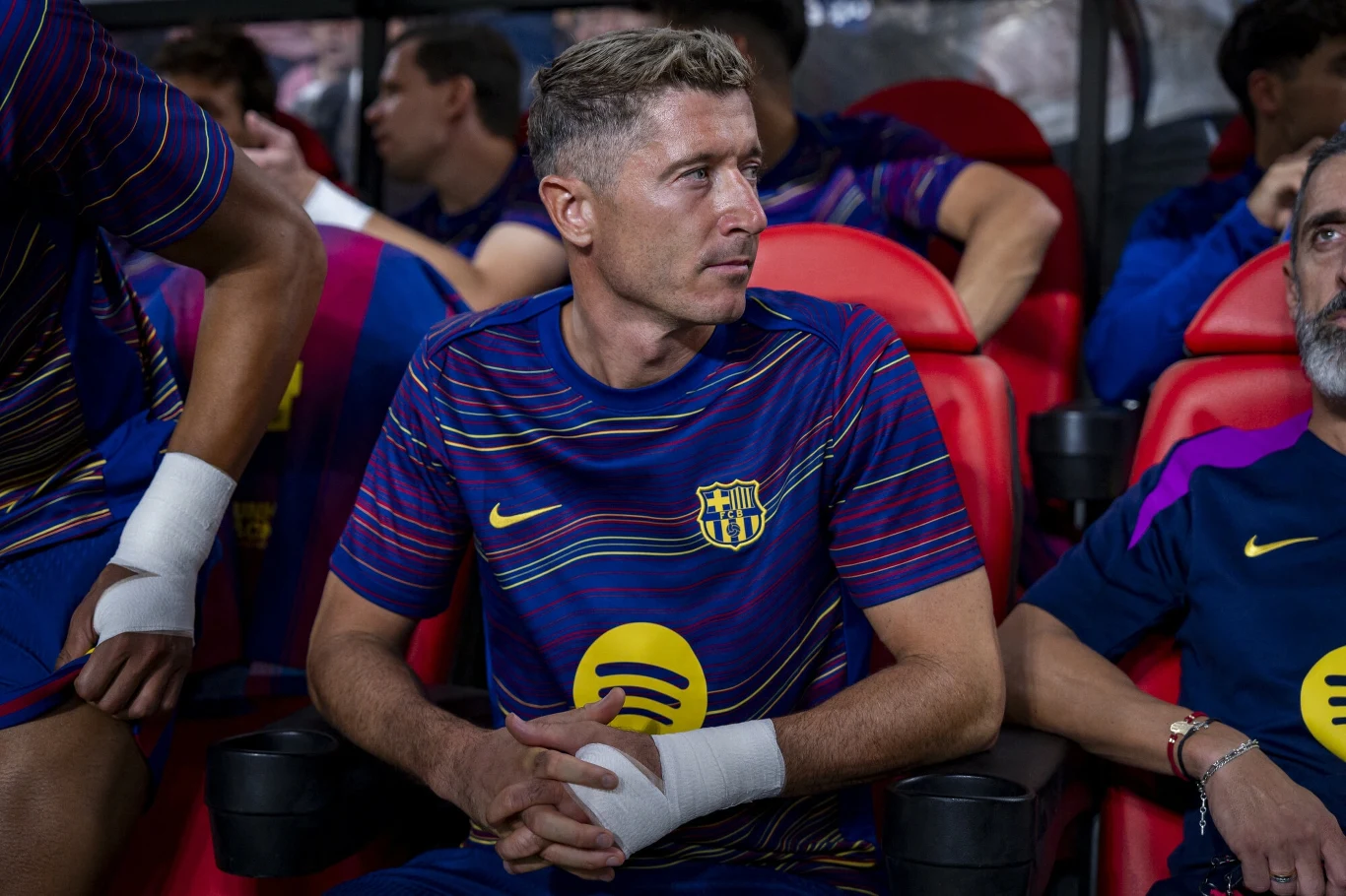 Robert Lewandowski Piłkarz w sportowym stroju drużyny FC Barcelona siedzi na ławce rezerwowych, otoczony przez innych członków zespołu, w tle widać kilku zawodników rozmawiających ze sobą.