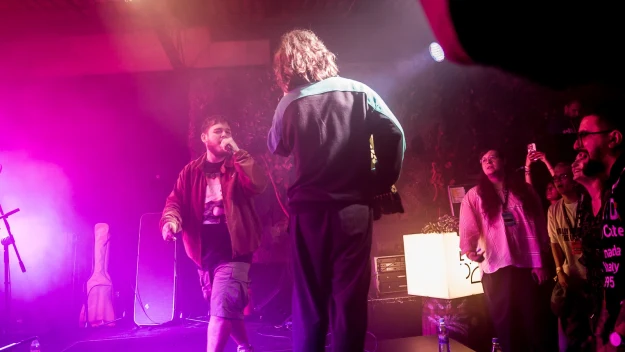 Dwóch artystów na scenie podczas koncertu, jeden stoi przodem i śpiewa do mikrofonu, drugi odwrócony tyłem, widoczna publiczność oraz kolorowe światła i efekty dymu.