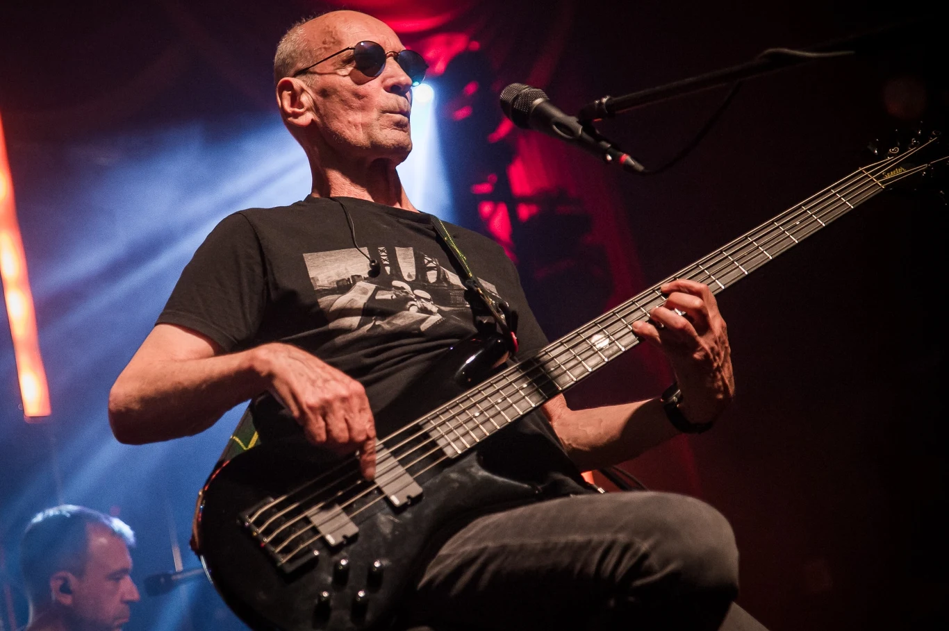 Great September 2025: Lech Janerka Starszy mężczyzna grający na gitarze basowej na scenie, ubrany w czarną koszulkę z nadrukiem, w okularach słonecznych, przy mikrofonie, z dynamicznym oświetleniem w tle dodającym koncertowego klimatu.