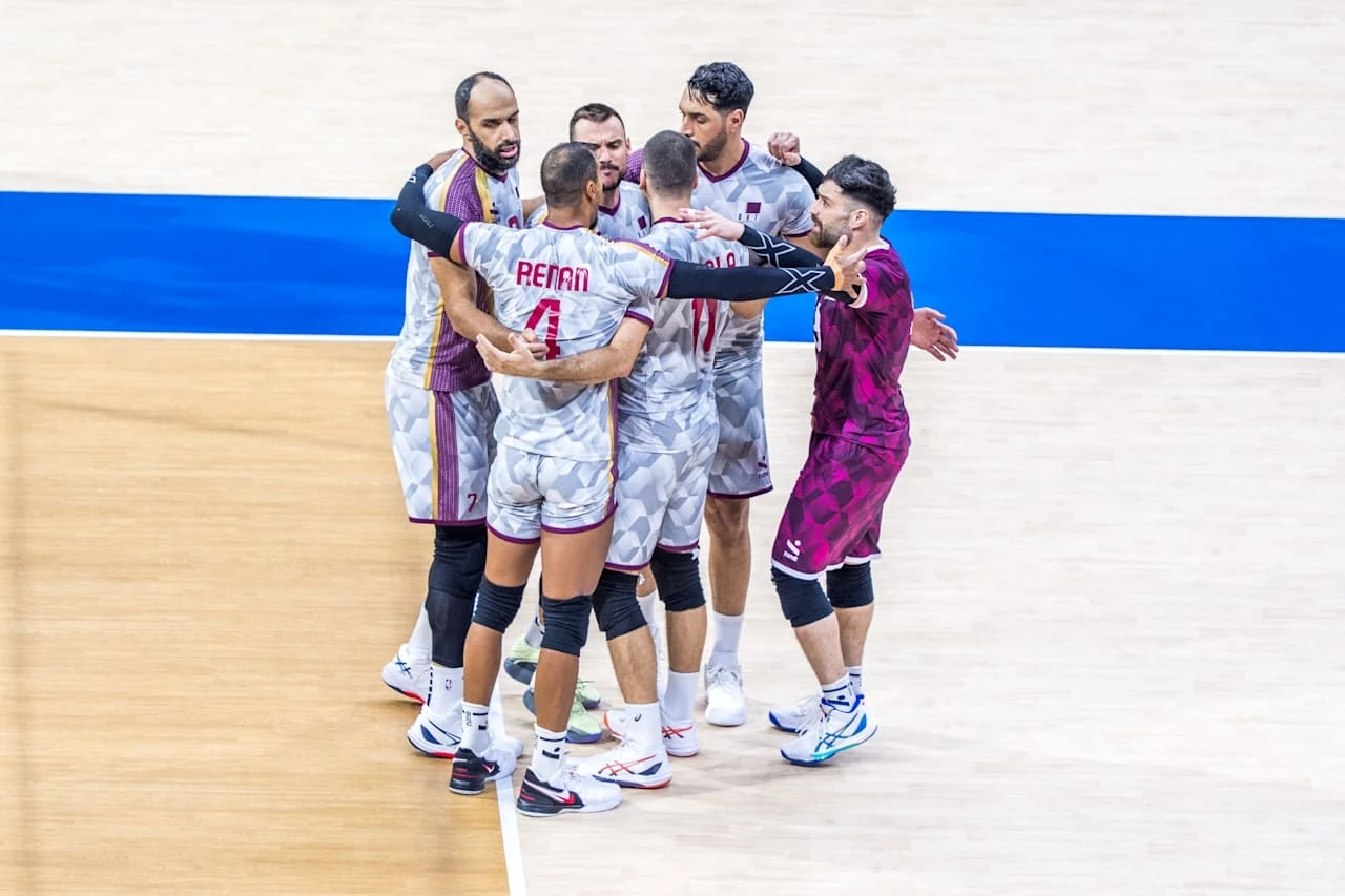 Katar na MŚ 2025 na Filipinach Grupa siatkarzy w sportowych strojach celebruje udaną akcję na boisku, otaczając się ramionami w geście radości i wsparcia drużynowego, na tle parkietu oraz niebieskiej linii granicznej boiska.
