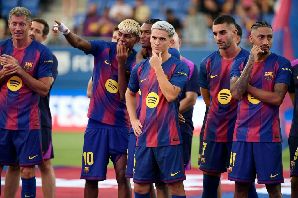 Grupa piłkarzy drużyny FC Barcelona w granatowo-bordowych strojach klubowych stoi razem na murawie stadionu, kilku zawodników prezentuje zamyślone lub rozbawione wyrazy twarzy, na strojach widoczne logo sponsora Spotify.