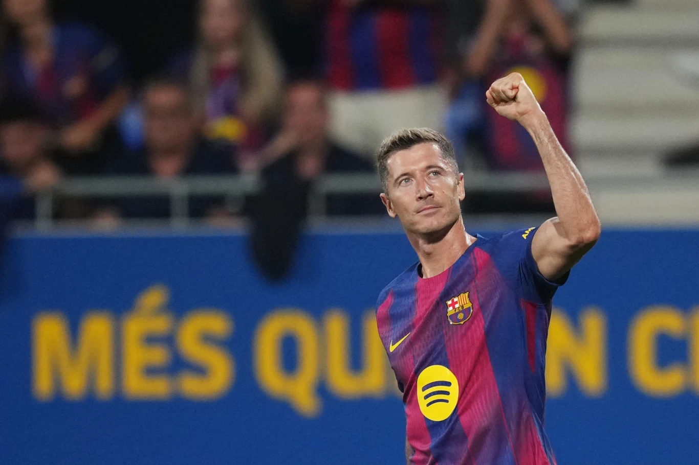 Piłkarz drużyny FC Barcelona w klubowej koszulce cieszy się po zdobyciu gola, unosząc zaciśniętą pięść w geście triumfu, w tle wyraźnie widoczna żółta tablica ze sloganem klubu oraz widownia.