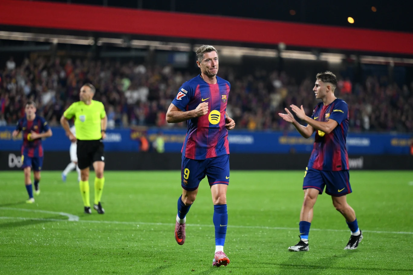 Piłkarze drużyny w granatowo-czerwonych strojach cieszą się po zdobyciu gola na tle pełnych trybun; w tle widoczny sędzia w jaskrawożółtym stroju oraz kibice na stadionie.