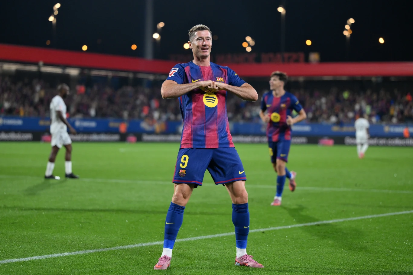 Piłkarz drużyny FC Barcelona w charakterystycznej koszulce wykonuje gest zwycięstwa na boisku podczas nocnego meczu, w tle widoczny tłum kibiców i pozostali zawodnicy.