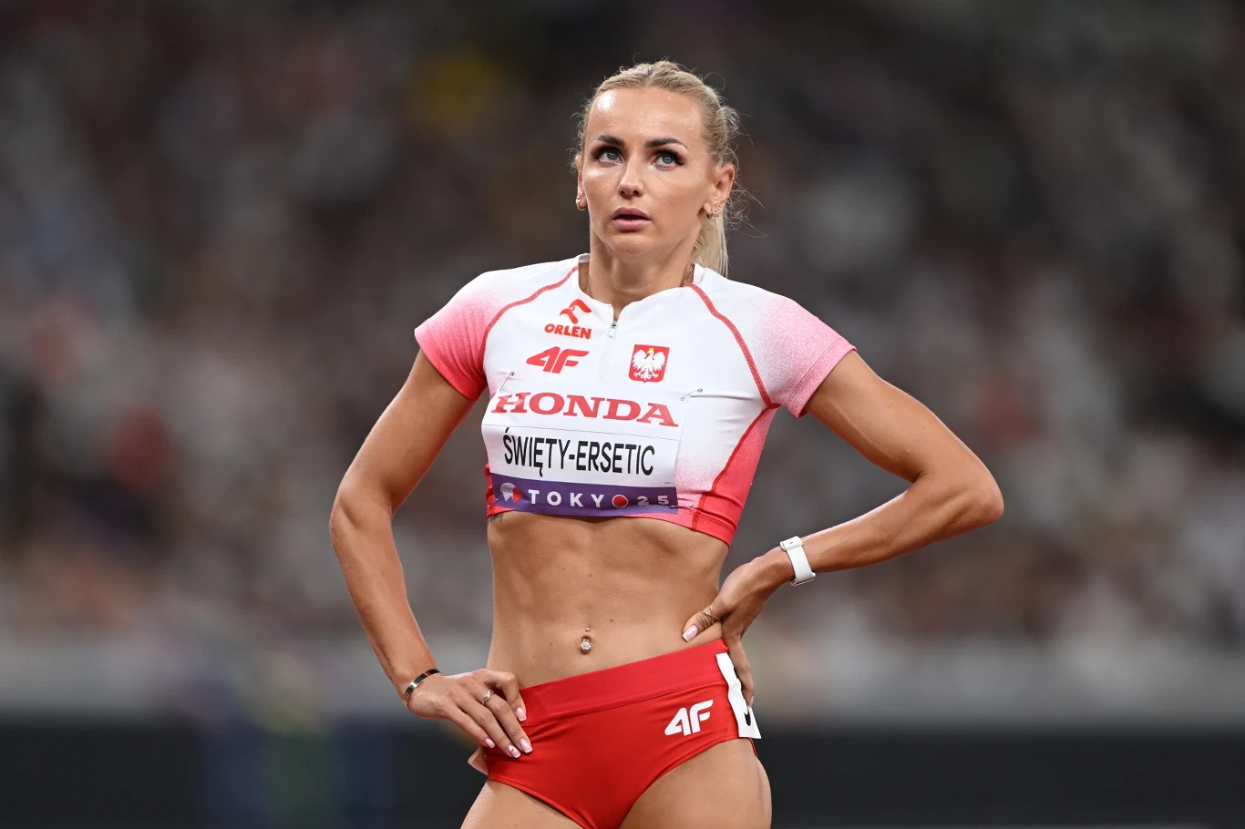 Justyna Święty-Ersetic Lekkoatletka w stroju reprezentacyjnym z napisem Polska i nazwiskiem na koszulce, w tle niewyraźne zarysy stadionu, sportowy wyraz koncentracji i gotowości do startu.