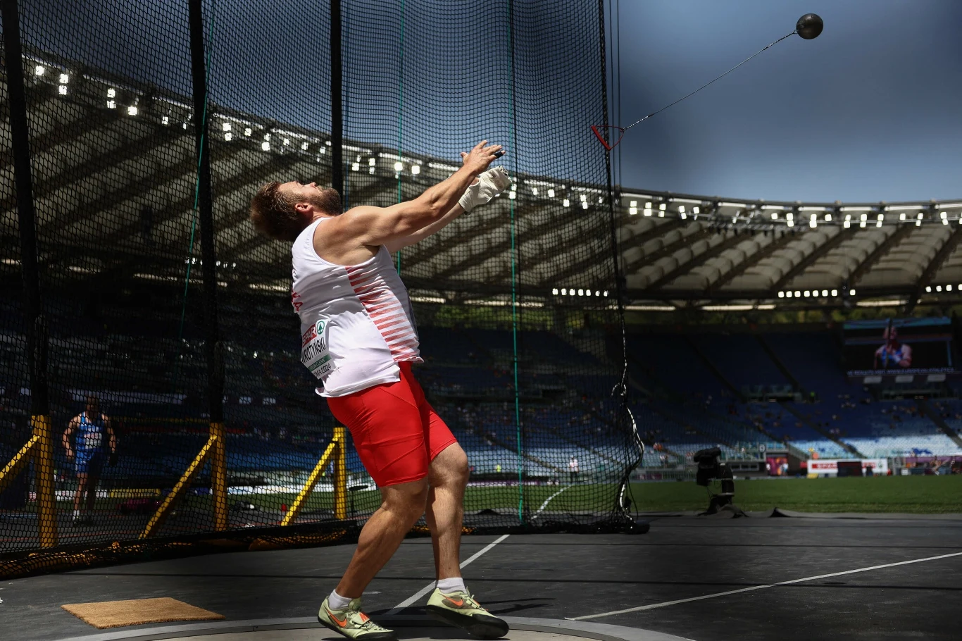 Marcin Wrotyński Lekkoatleta w trakcie rzutu młotem na stadionie, w tle trybuny i siatka ochronna. Zawodnik ubrany w biało-czerwony strój sportowy.