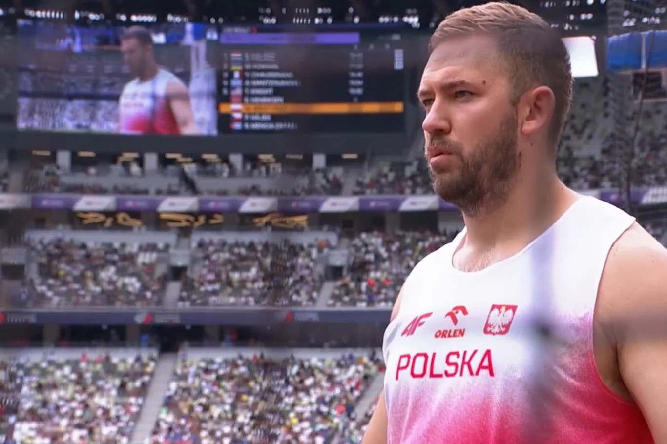 Marcin Wrotyński Lekkoatleta w stroju reprezentacji Polski podczas zawodów lekkoatletycznych na stadionie, w tle widoczna publiczność oraz tablica wyników.