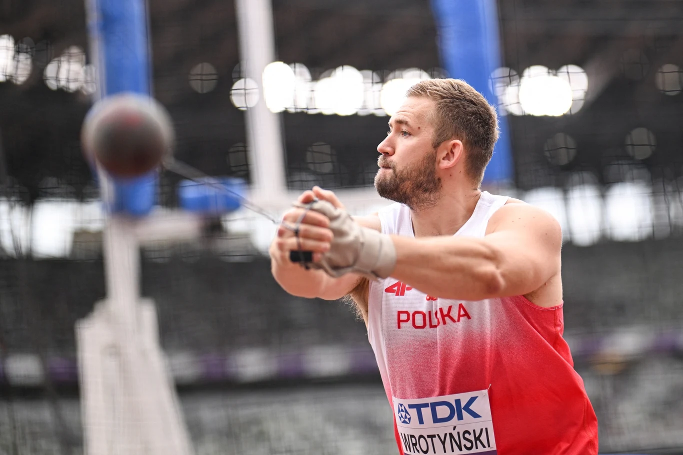 Marcin Wrotyński, Tokio 2025 Lekkoatleta w białoczerwonym stroju z napisem Polska wykonuje rzut młotem na stadionie lekkoatletycznym, w tle rozmyte trybuny i elementy infrastruktury sportowej.