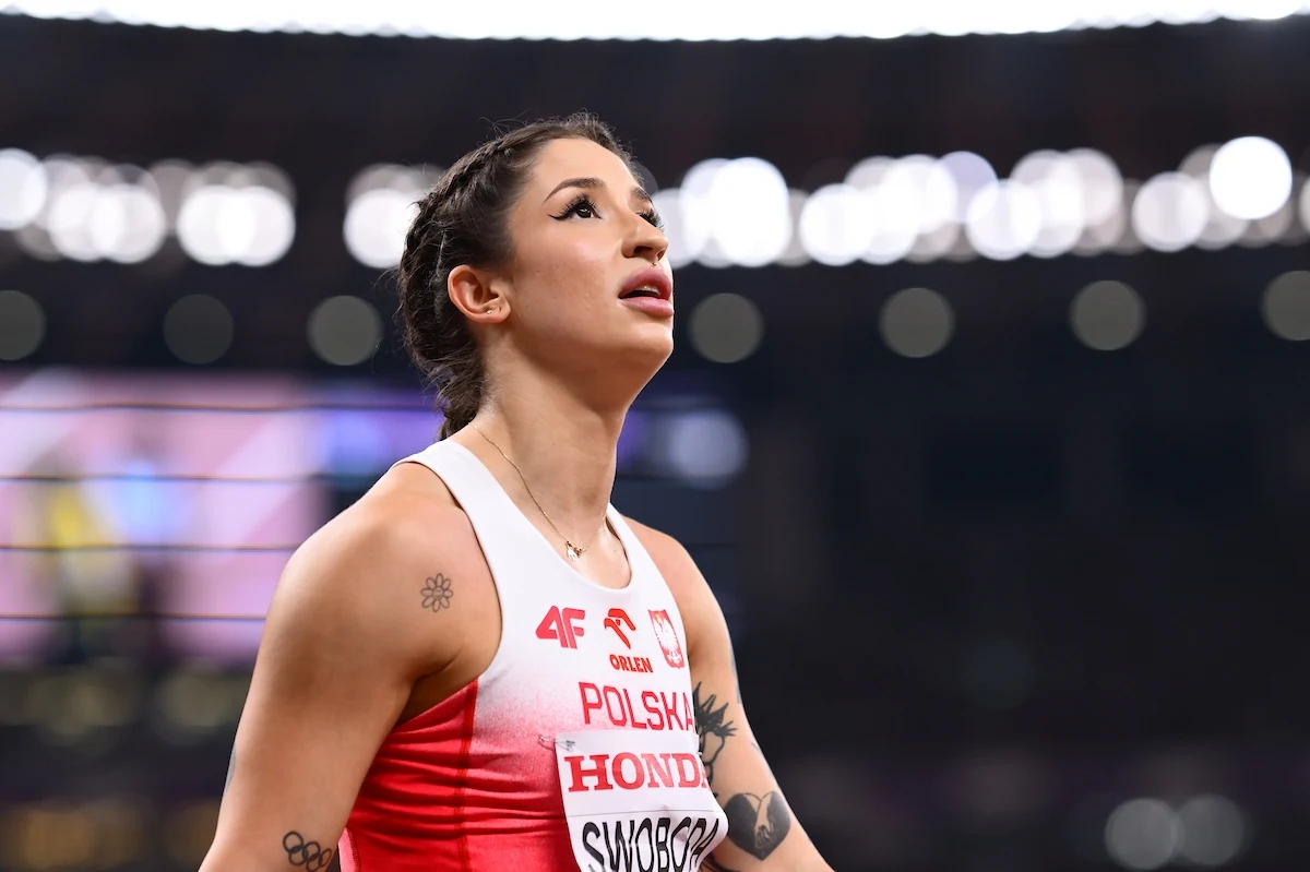 Lekkoatletka w biało-czerwonym stroju reprezentacji Polski z widocznym napisem na koszulce oraz numerem startowym, skupiona i patrząca w górę, w tle rozmazane światła stadionu.
