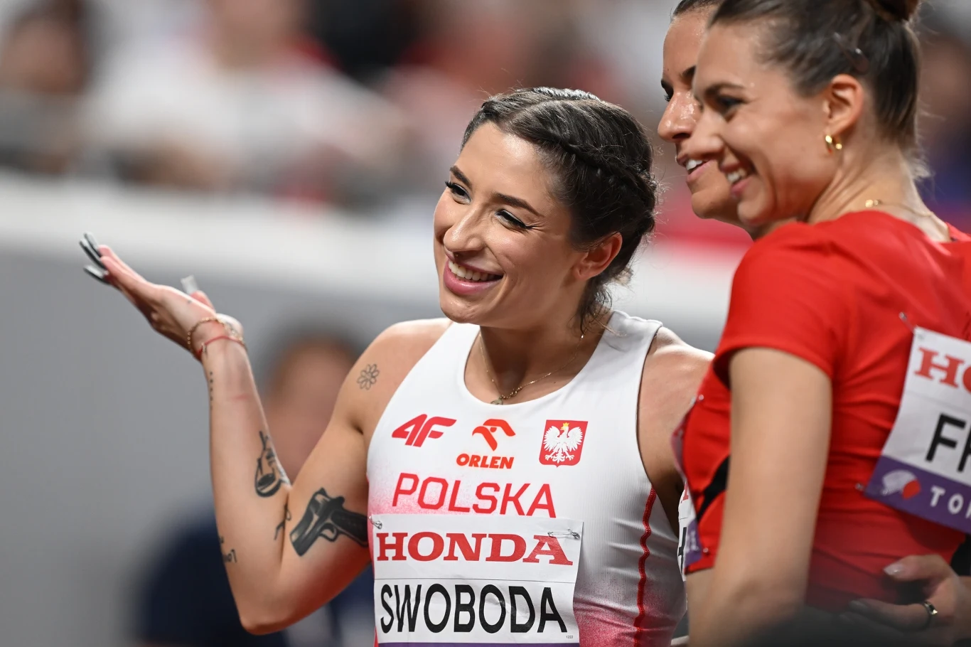 Uśmiechnięta lekkoatletka reprezentująca Polskę podczas zawodów sportowych, ubrana w biało-czerwony strój z logotypami sponsorów, w towarzystwie innej zawodniczki.