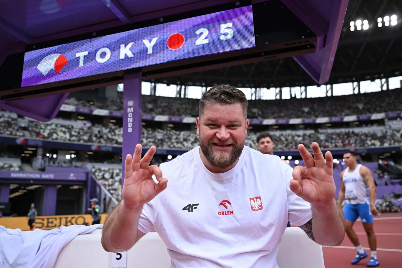 Uśmiechnięty mężczyzna w białej koszulce z polskim godłem siedzi na stadionie lekkoatletycznym, pokazując gest OK obiema rękami, w tle widoczny elektroniczny baner z napisem TOKYO 25 oraz inni zawodnicy.