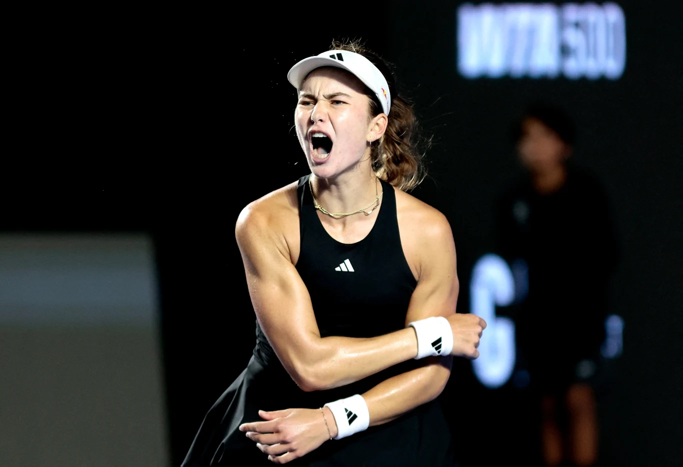 Tenisistka ubrana w czarny strój sportowy i białą opaskę na głowie z wyrażeniem silnych emocji na twarzy, ściska ramię po dynamicznym zagraniu na tle rozmazanego ciemnego tła z podświetlonym napisem.